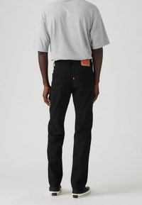 Vaqueros negros de corte recto, con bolsillos traseros visibles y una parche de cuero con la marca en la cintura. Combinados con una camiseta de manga corta gris.