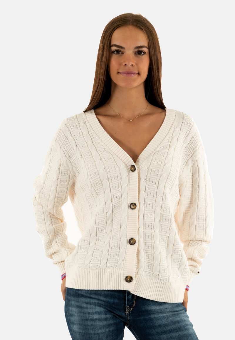 Giovane donna con lunghi capelli marroni che indossa un cardigan a costine color crema con bottoni e jeans blu, in piedi con le mani nelle tasche su uno sfondo bianco.