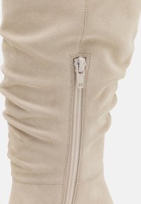 Béžový semišový svršek boty se vertikálním béžovým zipem a viditelným šitím na bílé pozadí.