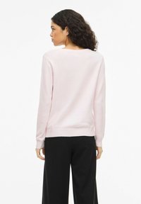 Femme aux cheveux bouclés et foncés, portant un pull léger rose pâle à manches longues et un pantalon noir à jambes larges, vue de dos.