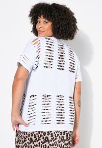 Valkoinen ylipitkä t-paita, jossa on useita leikattuja rakoja takana, paljastaen vastakkaisen mustan kerroksen alla. Lyhyet hihat.