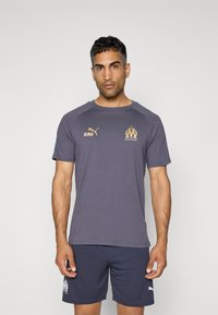 Grå T-shirt med blå accenter og gyldne logoer, der har rund hals og korte ærmer, parret med navy shorts med logo ved kanten.