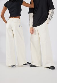 Pantalons larges blancs avec une poche arrière arborant un visage souriant bleu. Un t-shirt noir court et une chemise à manches longues superposée noir et blanche.