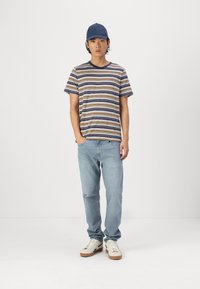 T-shirt à manches courtes rayé en navy, beige et marron, associé à un jean bleu clair et des baskets blanches avec des semelles en caoutchouc beige et des accents.