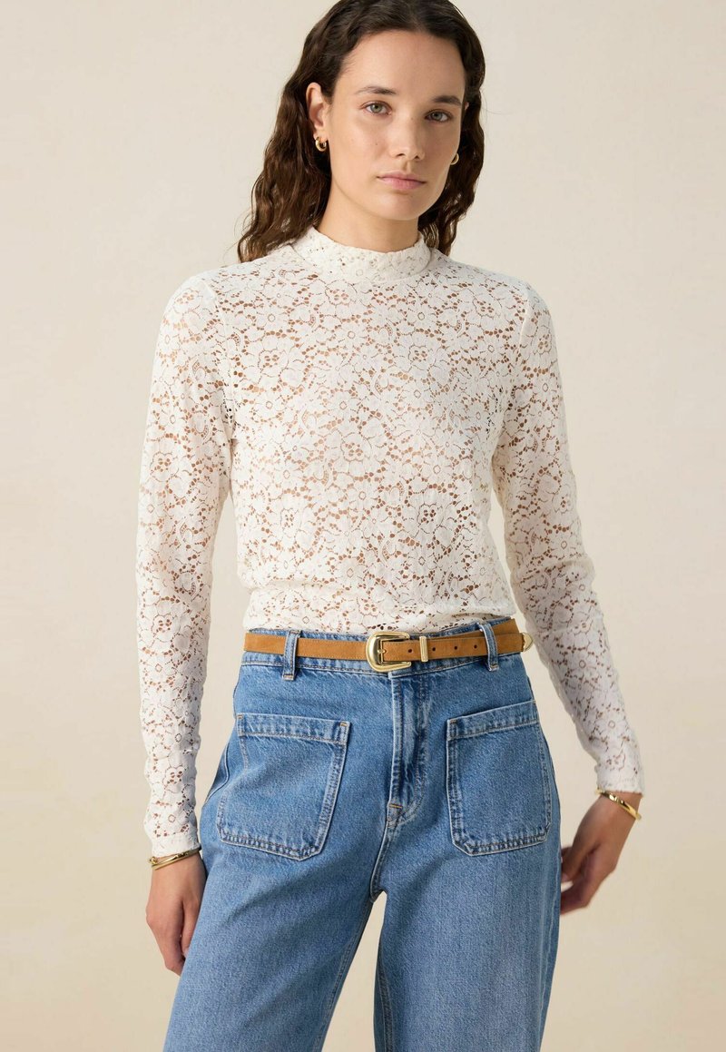 Spitzen-Longsleeve-Oberteil in Off-White, mit floralen Mustern und hohem Kragen, kombiniert mit hellblauen, hoch taillierten Jeans und einem braunen Gürtel.