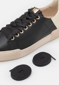 Baskets en cuir noir avec œillets dorés et semelle en caoutchouc beige. Comprend deux lacets en tissu noir, disposés en spirales à côté de la chaussure.