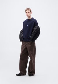 Μπλε πλεκτό πουλόβερ με ριμπ μανσέτες, συνδυασμένο με φαρδιά καφέ παντελόνια από βελούδο κοτλέ. Το μοντέλο κρατάει μια μαύρη μπουφάν-φουσκωτό, στέκεται μπροστά σε ανοιχτό φόντο.