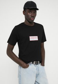 Μαύρο βαμβακερό μπλουζάκι με ορθογώνια λευκή ετικέτα με κόκκινο κείμενο. Συνδυασμένο με ανοιχτού μπλε τζιν παντελόνι και μαύρο καπέλο.