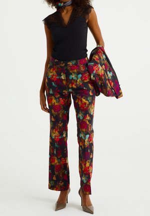 Femme portant un haut noir sans manches avec bordure en dentelle, pantalon fleuri multicolore, tenant une veste assortie à motif floral et chaussée de talons beiges.