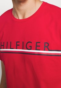 Hombre vistiendo una camiseta roja con rayas negras y blancas y el texto "HILFIGER" en el pecho.