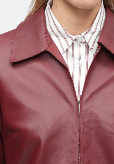 Κοντινό πλάνο ενός ατόμου που φορά ένα μπουφάν από μπορντό δέρμα πάνω από ένα λευκό πουκάμισο με κάθετες κόκκινες ρίγες και γιακά με κουμπιά.