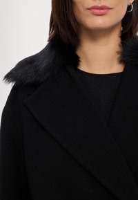 Femme portant un manteau noir à large col avec une bordure en fausse fourrure noire, associé à un haut noir texturé et une boucle d'oreille argentée en forme d'anneau.