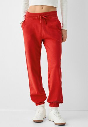 Person iført røde joggingbukser med snøre og elastiske manchetter, hvide sneakers og en hvid cropped langærmet top, stående mod en ensfarvet baggrund.