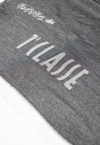 Sciarpa grigia con testo bianco "T'ILLASSE," bordi frangiati e una texture trasparente, che mostra un motivo sottile nel tessuto.