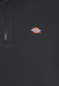 Black zip-up sweatshirt featuring a woven label with a multicolored circular logo that includes the brand name "Dickies." Smooth fabric texture.