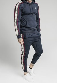 Marinblå joggingdräkt med vita och röda sidostreck, bestående av en huva sweatshirt och avsmalnande byxor. Tillverkad av slätt tyg; inkluderar logodetaljer.