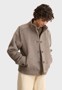 Brown and beige checked jacket with a high collar, front buttons, and side pockets. Fabric appears textured and warm.