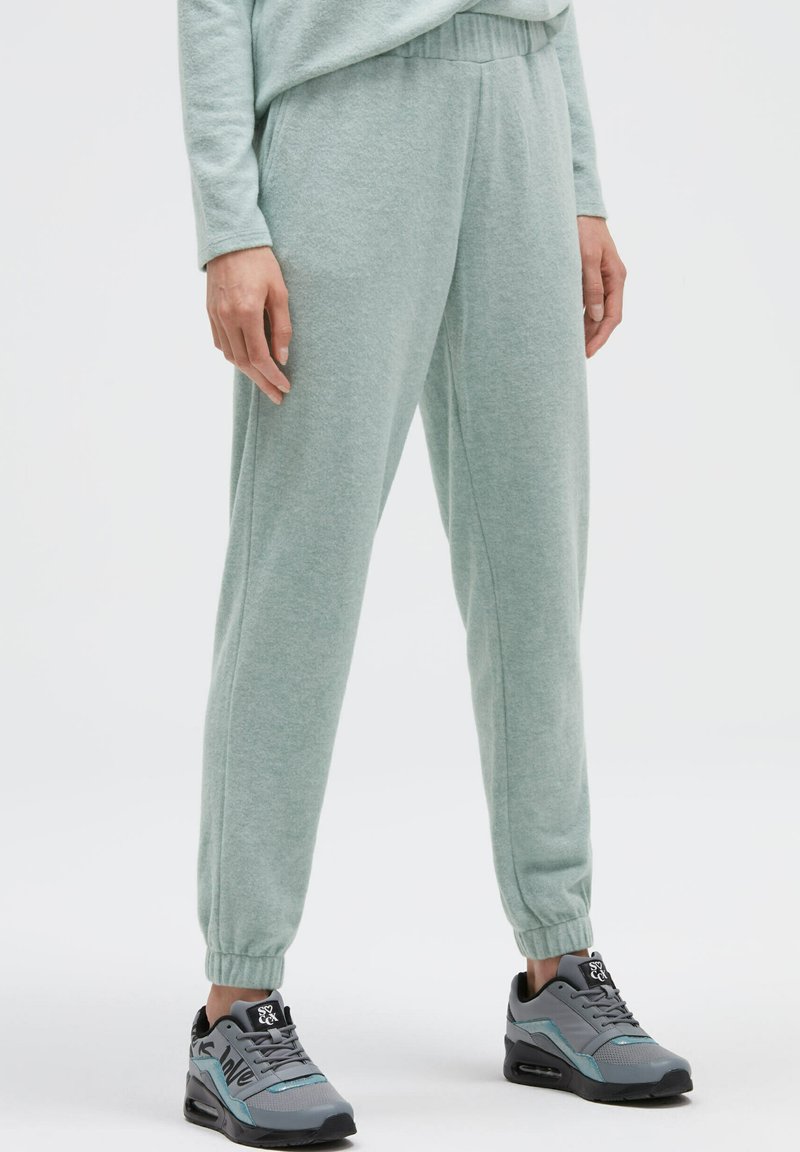 Hellgrüne, weiche Stoff-Jogginghosen mit schmal zulaufendem Bein, elastischem Bund und gerippten Bündchen, kombiniert mit grauen Sportschuhen mit türkisfarbenen Akzenten.