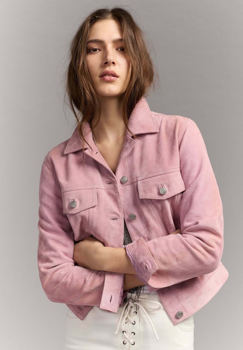 Donna con capelli castani lunghi indossa una giacca di camoscio rosa chiaro con bottoni d'argento, con le braccia incrociate sopra pantaloni bianchi con lacci, su sfondo grigio.