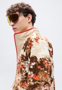 Φλις μπουφάν σε μπεζ απόχρωση με λουλουδάτο μοτίβο σε καφέ και πορτοκαλί, με ψηλό γιακά και κόκκινο φερμουάρ. Φορεμένο με σπορ ηλιακά γυαλιά με κίτρινο φιμέ φακό.