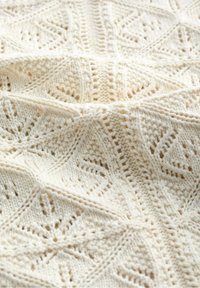 Kremowy koc dziany z misternymi wzorami koronkowymi, zawierający diamentowe kształty i fakturowane ściegi, tworzący miękki i przytulny wygląd.