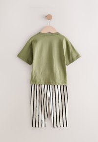 Zöld, rövid ujjú pamut póló csíkos krémszínű és fekete nadrág fölött lóg. Egyszerű dizájn, sima textúra és laza stílus.