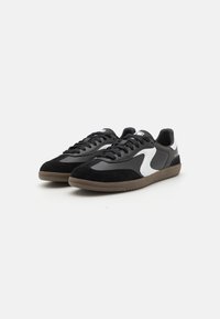 Sorte og hvite sneakers med en overdel i semsket skinn og syntetisk materiale, med en avrundet tå, flate lisser og en teksturert brun gummisåle.