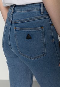 Jeans de ganga azuis de cintura alta com um acabamento desbotado, apresentando cinco bolsos e um logo triangular preto no bolso de trás.