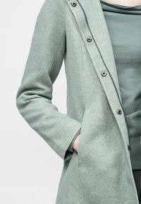 Lyst grønn teksturert kåpe med glidelås og trykknapper, med en skreddersydd passform og sidelommer, lagd over en matchende topp.