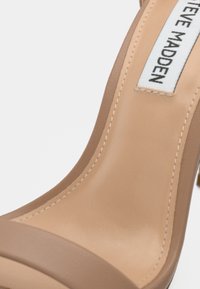 Beige korkokenkä, jossa on sileä synteettinen päällinen, leveä etuolka ja merkitty kangaslabeli. Reunassa on ommeltuja yksityiskohtia.