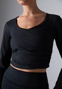 Žena nosi uski crni dugosleevi crop top s omotanim dekolteom i visokim crnim donjim dijelovima uz neutralnu pozadinu.