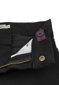 Mustat farkut, joissa on kultainen vetskari, punainen ja sininen raidoitettu kangas yksityiskohtana napissa, sekä valkoinen vuori näkyvissä taskussa.