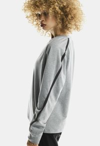 Woman with curly blonde hair wearing a grey long-sleeve shirt with black stripes along the sleeves standing sideways against white background.