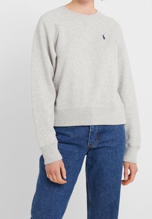 Persoon draagt een lichtgrijze trui met lange mouwen en een klein marineblauw logo op de borst, gecombineerd met blauwe spijkerbroek, staand tegen een effen achtergrond.