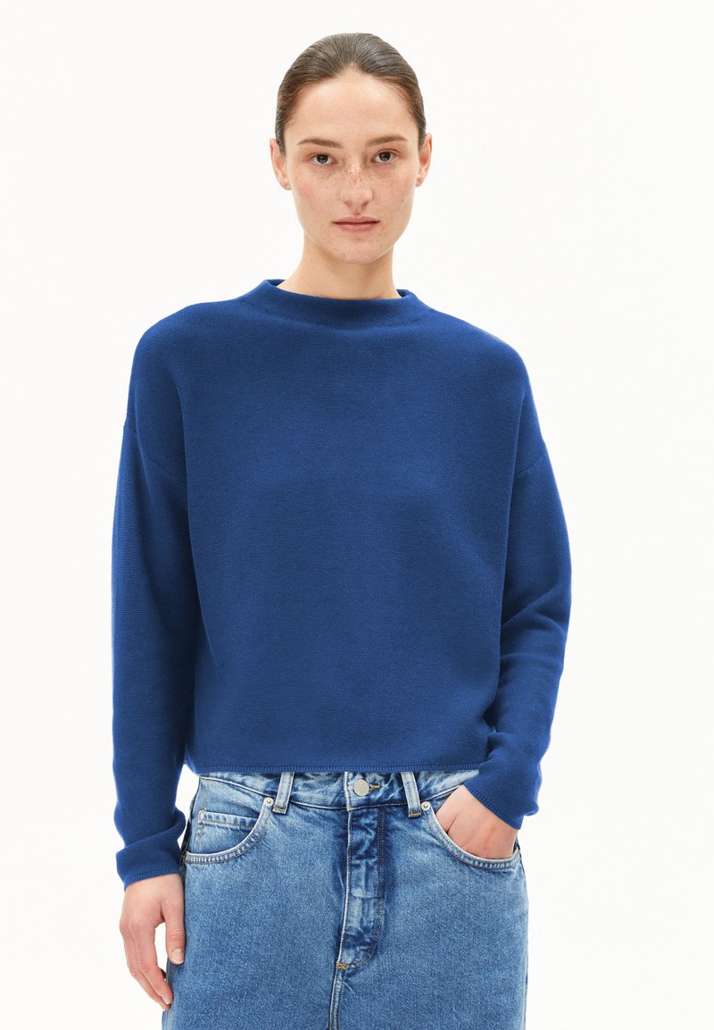 Blauer gestrickter Pullover mit hohen Kragen, überschnittenen Schultern und gerippten Bündchen, kombiniert mit hellblauen Jeansshorts mit seitlichen Taschen.