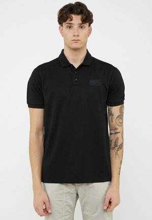 Joven con cabello rizado que lleva una camiseta polo negra y pantalones beige, mostrando tatuajes en su brazo derecho contra un fondo liso.