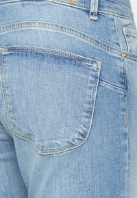 Jeans de ganga azul claro com um bolso traseiro com borda costurada e um detalhe em botão dourado. Textura suave e design clássico de cinco bolsos.