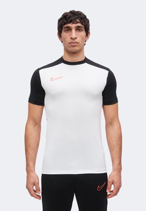 Junger Mann trägt ein enganliegendes weiß-schwarzes Sportshirt mit einem orangefarbenen Nike-Logo, steht vor einem schlichten hellen Hintergrund.