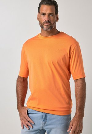 Mann mit kurzen dunklen Haaren und Bart trägt ein oranges kurzärmeliges Hemd und hellblaue Jeans, steht mit einer Hand in der Tasche vor einfarbigem Hintergrund.