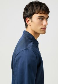 Camisa azul marino de tejido suave con cuello abotonado, que muestra una vista lateral que resalta las costuras de los hombros y los detalles del corte ajustado.