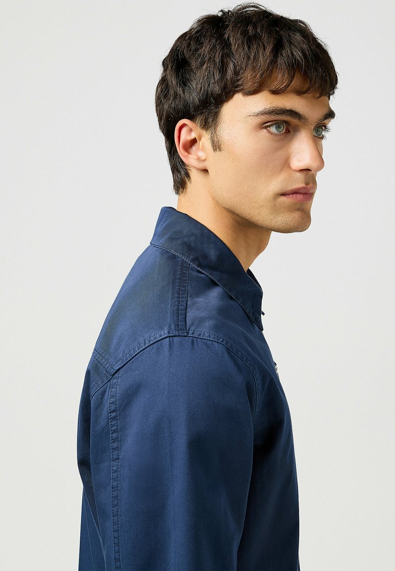 Granatowa koszula z gładkiego materiału z kołnierzykiem na guziki, z widokiem z boku uwypuklającym szwy na ramionach oraz szczegóły w dopasowanym kroju.