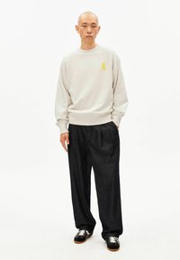 Light gray sweatshirt with a small yellow logo, paired with dark blue pleated trousers and black sneakers with white laces.
