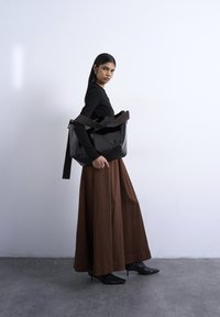 Γυναίκα με μαύρο τοπ, μακριά καφέ φούστα και μαύρες μπότες με τακούνι, κρατώντας μεγάλη μαύρη τσάντα στον ώμο, που στέκεται απέναντι σε απλό τοίχο.