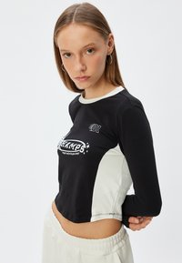 Langslibbige crop top van zwarte katoen met crèmekleurige zijkanten. Voorzien van een grafische logo-print en geribde ronde hals. Soepele textuur.