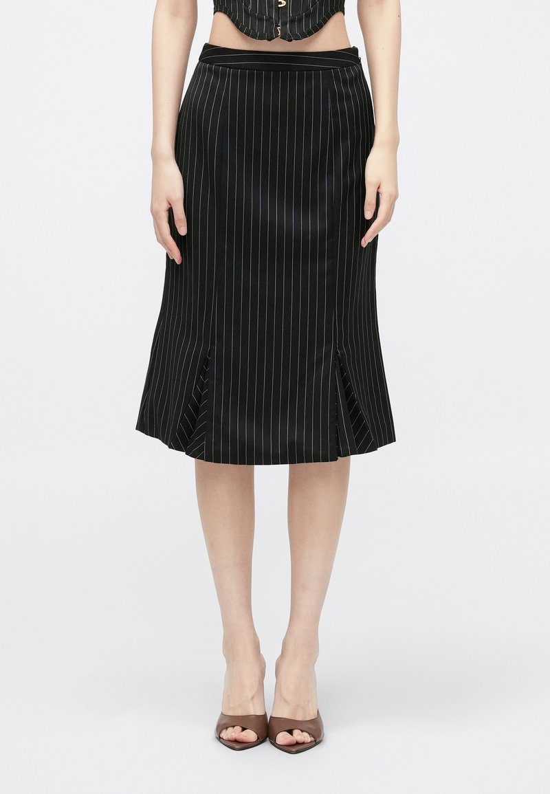 Juoda midi sijonas su juostelėmis ir išplatintu siūlu, turintis šonines skylutes ir lygaus, satino tipo tekstūrą. P wornas su rudiem atvirais aukštakulniais.