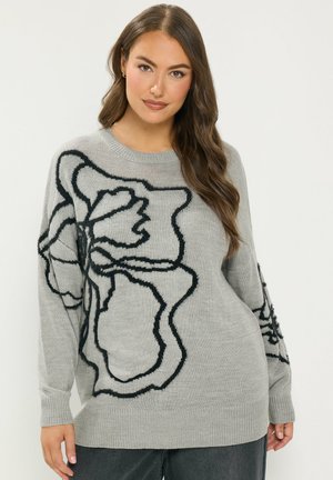 Donna che indossa un maglione grigio ampio con grandi motivi floreali astratti neri, abbinato a pantaloni scuri, in piedi contro uno sfondo semplice.