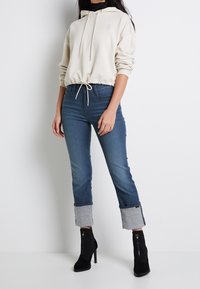 Krämfärgad kort hoodie med justerbar dragsko, tillsammans med ljusblå jeans med uppvikta fållar och svarta högklackade ankelstövlar.