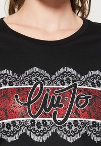 Schwarzes T-Shirt mit rundem Ausschnitt, das einen roten und weißen Mittelstreifen hat. Verz decorated mit einem Spitzenmuster und glitzerndem "Liu Jo"-Text.
