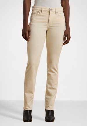 Nainen, joka käyttää beigen värisiä slim fit -housuja ja mustia nilkkureita, seisoo yksivärisen valkoisen taustan edessä.