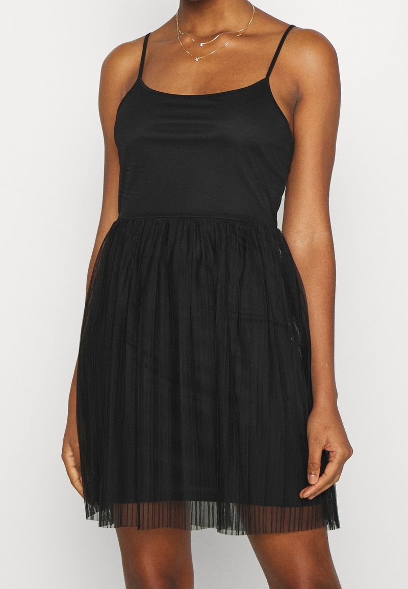 Kvinna klädd i en ärmlös svart klänning med tunna axelband och en plisserad kjol, prydnad med två eleganta guldsmycken.